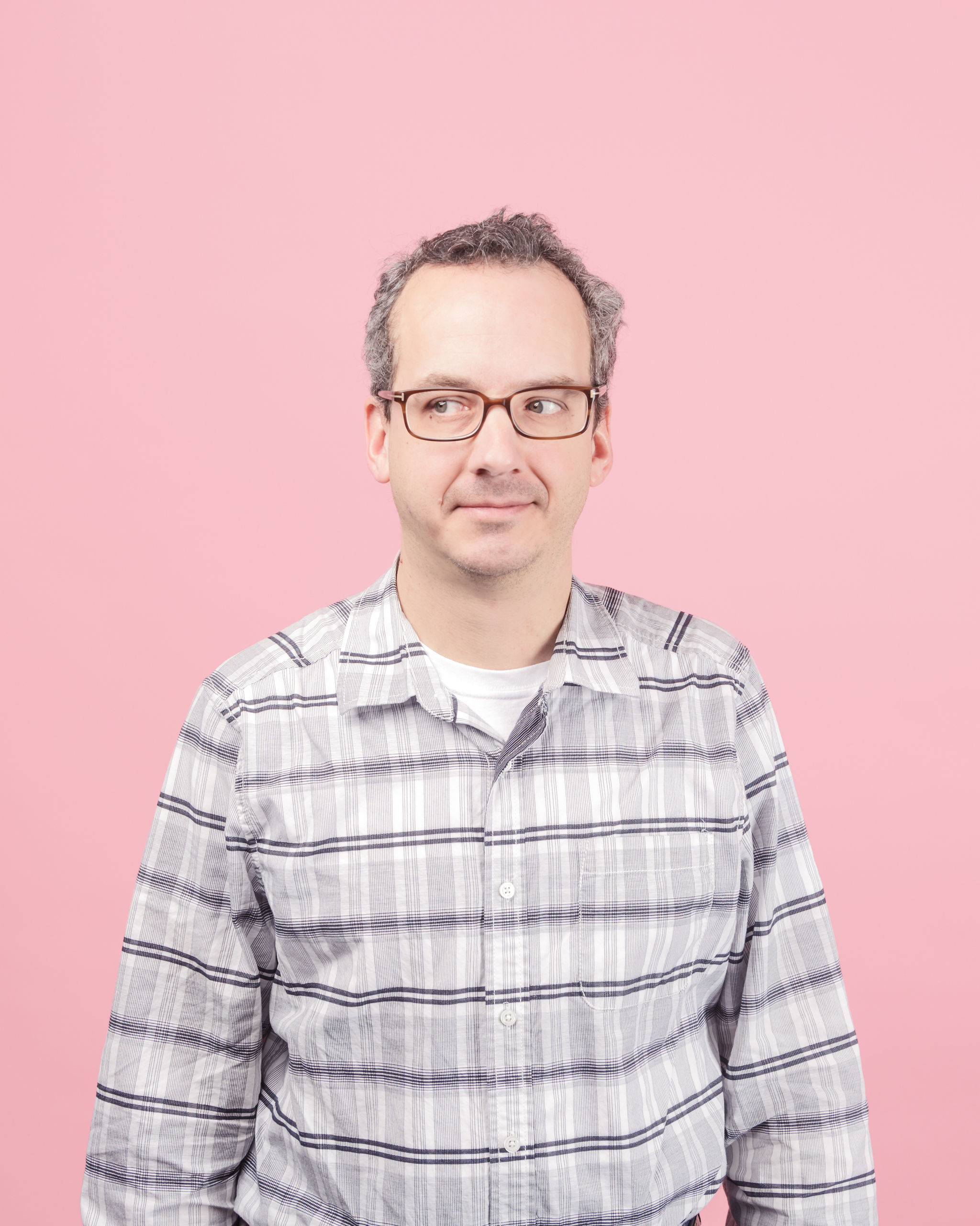 Headshot photograph of John Dunlevy facing forward with his eyes looking off to the right—wearing glasses and a button down black and white plaid pocket shirt while standing in front of a pink background