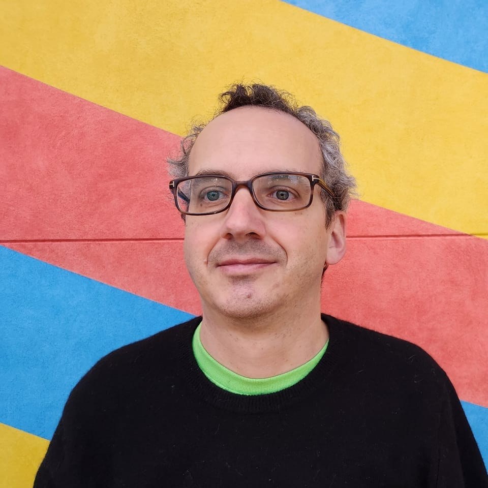 Medium-close headshot photograph of John Dunlevy wearing glasses and a black sweater over a green T-shirt in front of wall mural, diagonal blue, gold, and red stripes visible here